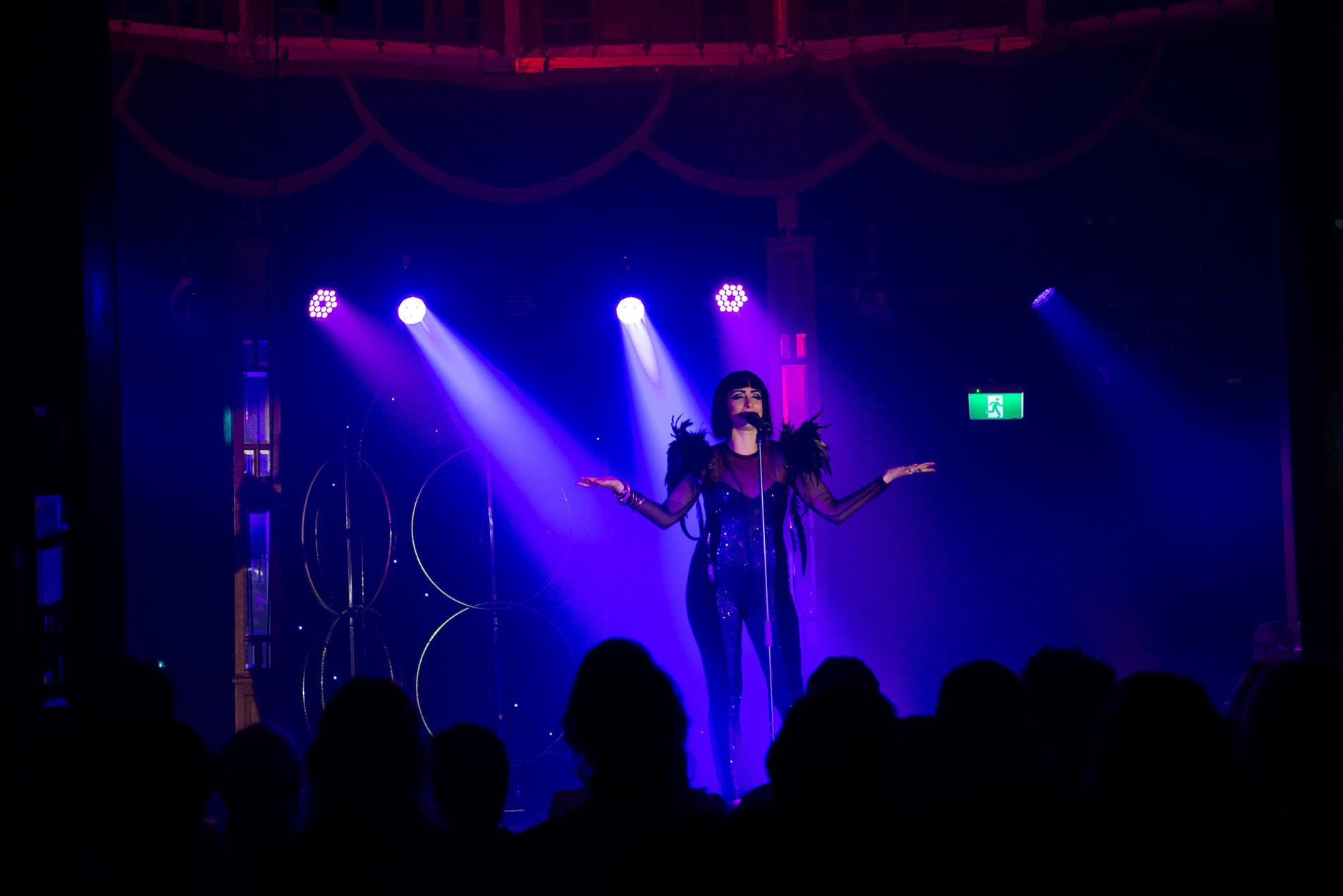 Performer in a black body suit with outstretched arms singing on stage.