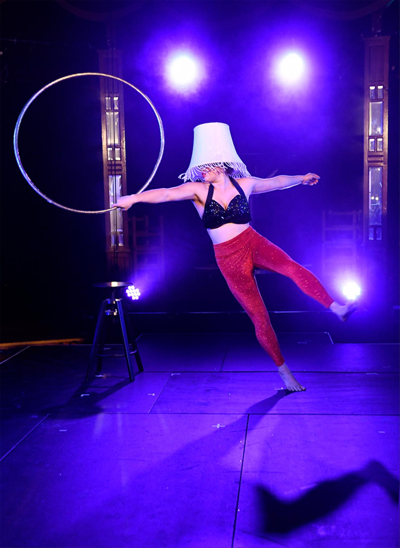 Woman gracefully dances with a hoola hoop, balancing a lamp shade on her head.