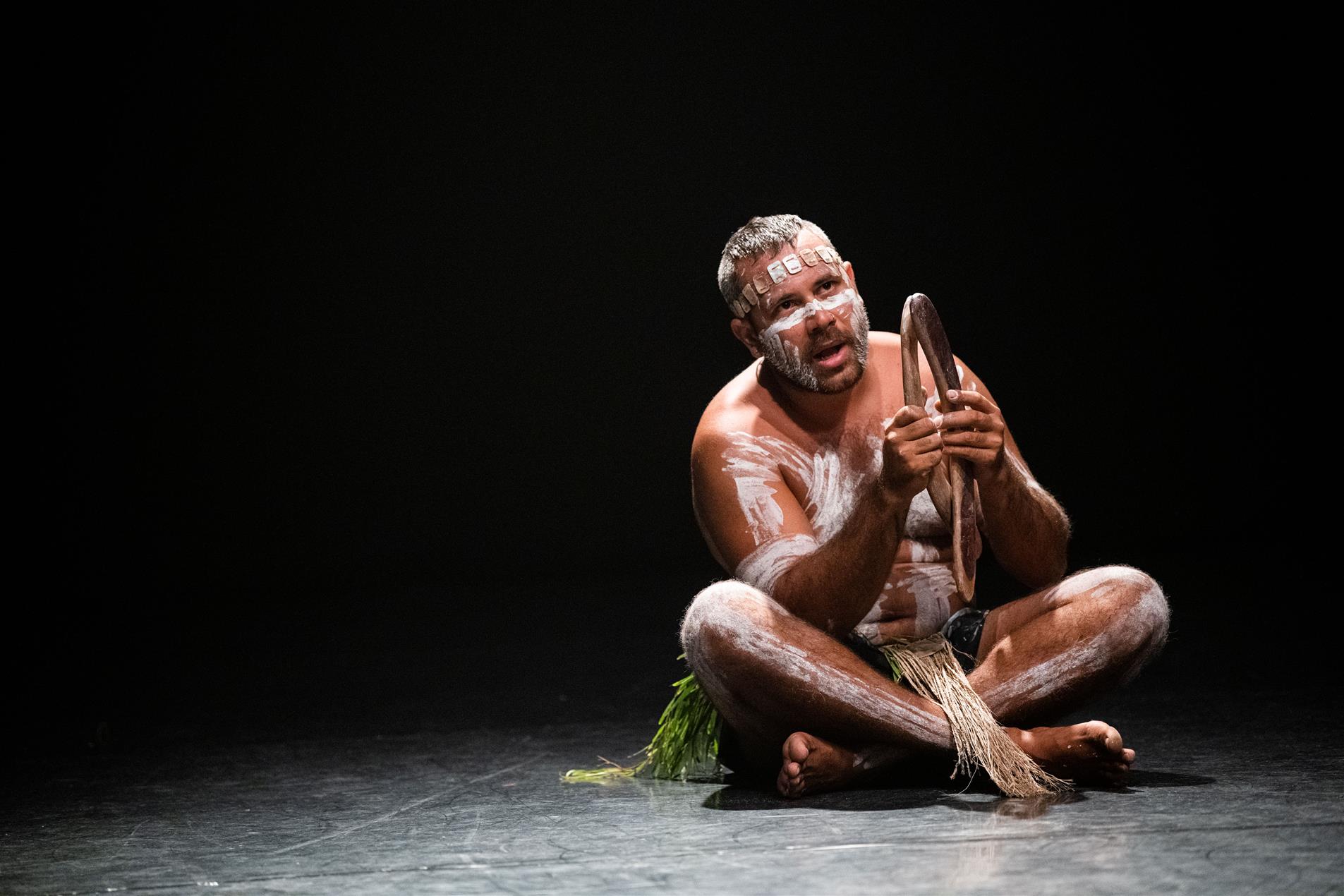 A man covered in white paint sitting on a stage performing with wooden sticks.