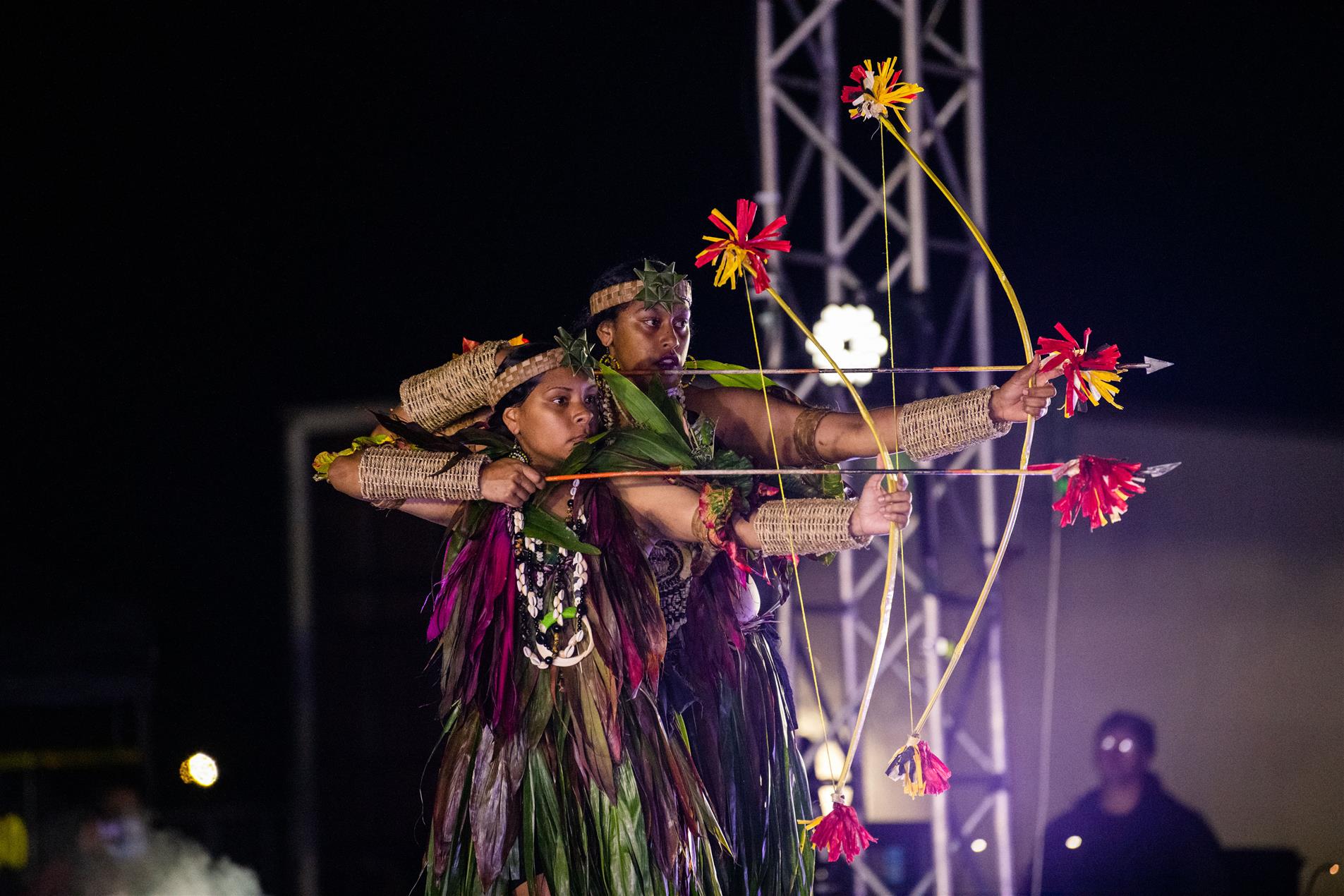 Two female performers wearing cultural attire holding bow and arrows.