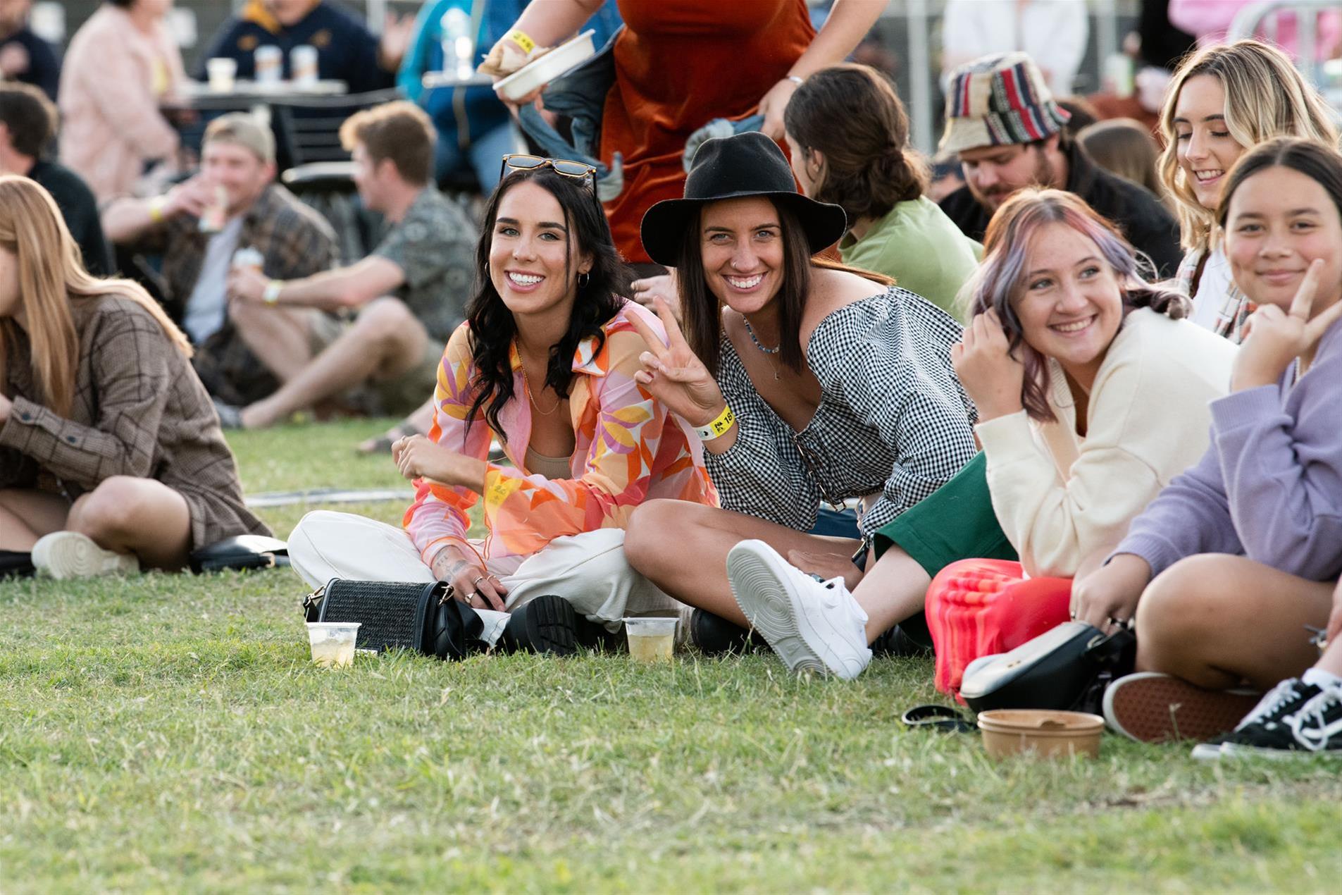 A group of females sitting on the grass at a music festival.