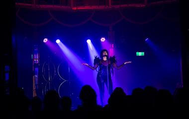 Performer in a black body suit with outstretched arms singing on stage.