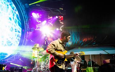 Three artists performing on stage. One female playing the piano, one female playing the drums and one male playing the guitar.